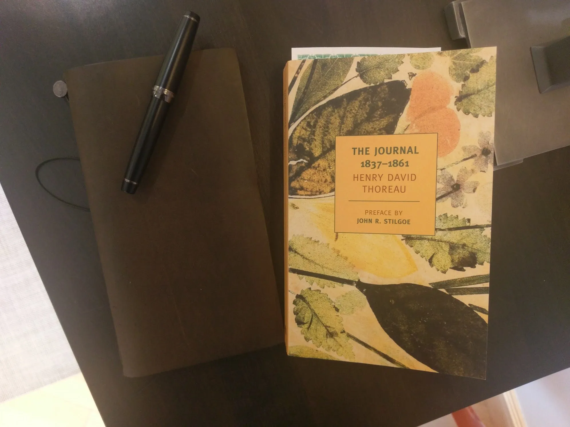 A black fountain pen resting on a brown leather journal beside a paperback of Henry David Thoreau's The Journal, 1837–1861.