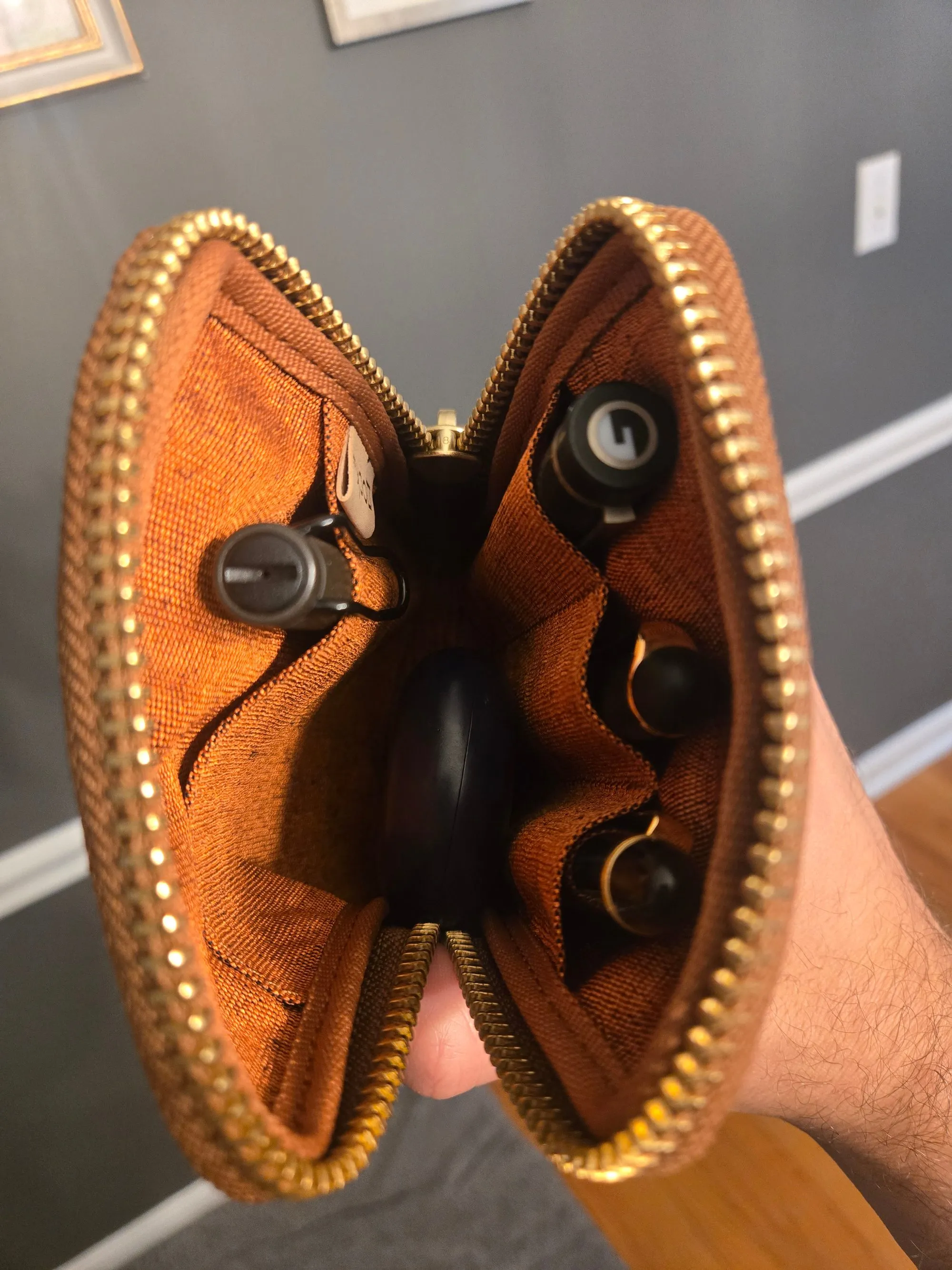 The open pen case showing three fountain pens in slots along the top, a black case tucked into the central compartment, and a rollerball pen clipped into the lower pocket, against rust-colored woven fabric.